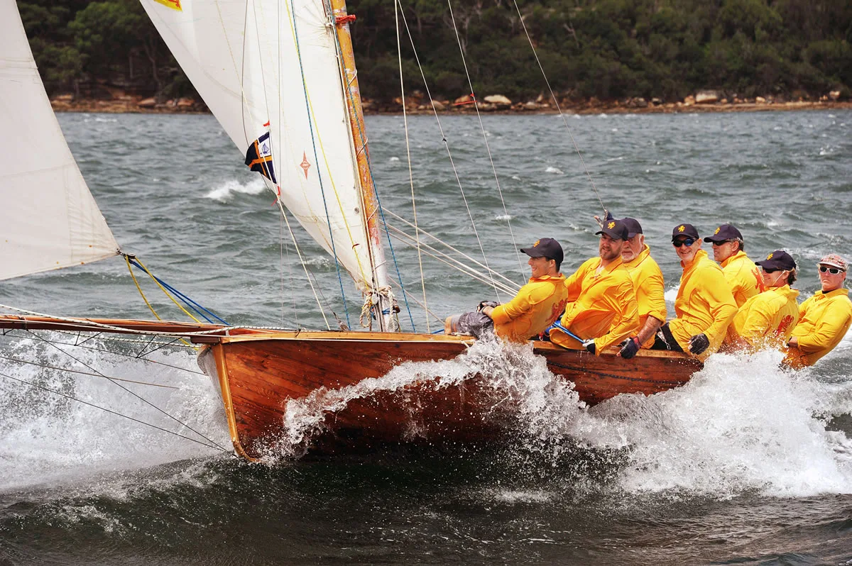 Crew in golden-yellow smocks hiked out as spray bursts over the bow of a varnished timber skiff